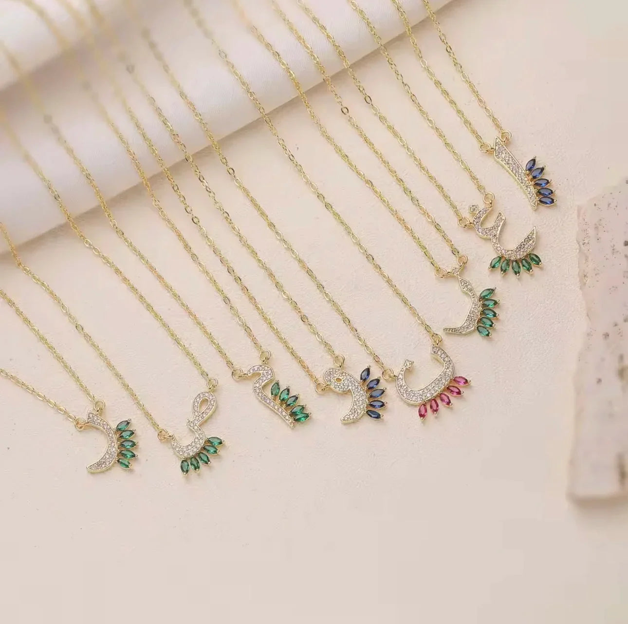 A set of gold-tone necklaces with peacock pendant charms, some adorned with clear rhinestones and others featuring green, blue, and pink gemstone accents, arranged on a light beige surface.